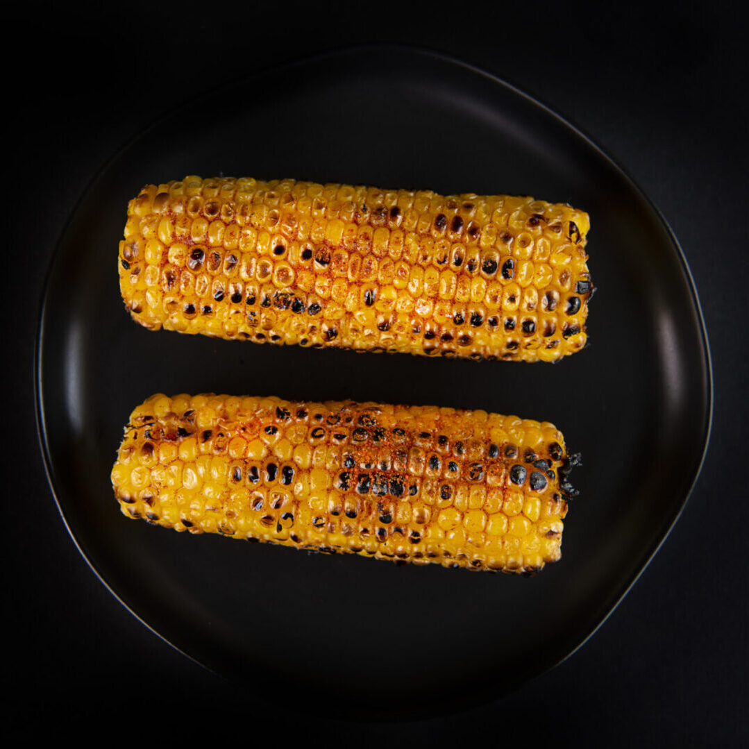 Two grilled corn on the cob sitting on a black plate.